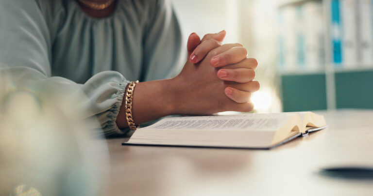 Woman wearing light blue top with hands crossed above an open bible
